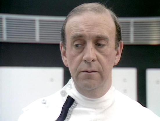 Claude 3.7 describes the image as: "The image shows a person in what appears to be a clinical or technical setting. They're wearing a distinctive white uniform or coat with some black detailing at the collar area. The background features what looks like control panels or display screens with a modern, futuristic aesthetic typical of science fiction productions from that era.

The setting suggests this might be from a scene in a medical bay, laboratory, or command center. The stark lighting and …