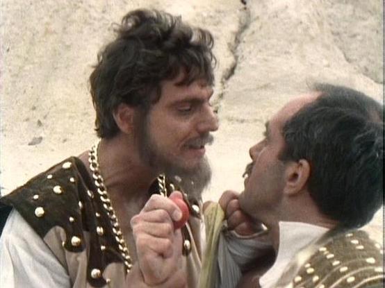 Claude Sonnet 4.0 describes the image as: "I can see this appears to be from a science fiction television production, showing what looks like a dramatic confrontation scene between two characters in ornate, futuristic costumes with metallic studs and decorative elements. The setting appears to be against a rough stone or cave-like background. The scene has the distinctive visual style of British science fiction television from the late 1970s or early 1980s, with elaborate costume design typical…