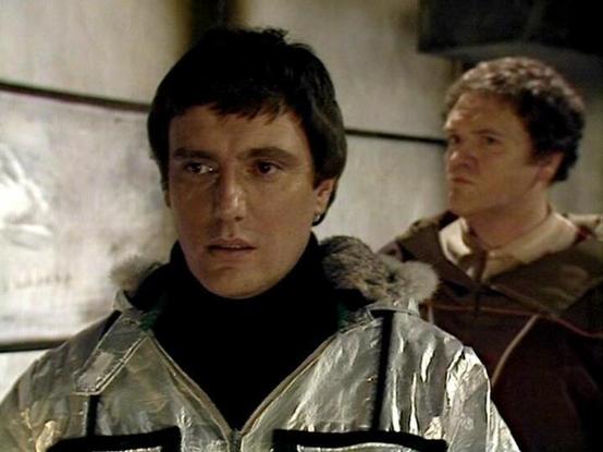 Claude 3.7 describes the image as: "This image appears to be from a science fiction television series from the late 1970s or early 1980s. The scene shows two individuals in what looks like a spacecraft or futuristic setting. In the foreground, a person is wearing a distinctive silver jacket with fur trim around the collar and a black turtleneck underneath, giving them a somewhat militaristic or space-age appearance. In the background, another person is partially visible wearing what appears to …