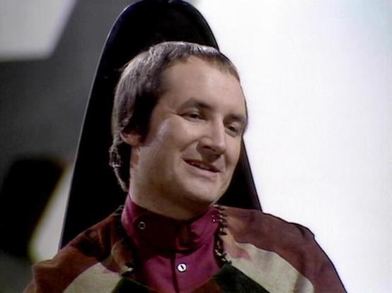 Claude 3.7 describes the image as: "This image appears to be from a science fiction television series from the late 1970s or early 1980s. The scene shows a person with short hair wearing a distinctive burgundy and brown patterned outfit with a high collar. They have a slight smile and appear to be in what looks like a futuristic spacecraft or space station setting, indicated by the sleek, minimalist background and what seems to be a chair or control station visible behind them.

The costume des…