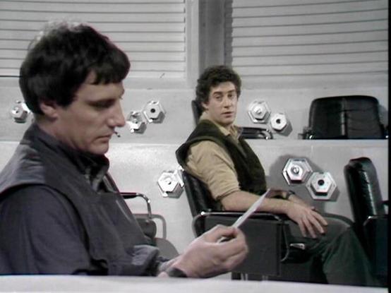 Claude Sonnet 4.0 describes the image as: "This scene takes place in what appears to be a sterile, white medical or detention facility. Two men are seated in the clinical environment - one in the foreground wearing dark clothing and looking downward with a troubled expression, while another sits in the background wearing lighter colored clothing with a vest. The setting has a cold, institutional feel with white walls and surfaces, along with hexagonal control panels or monitoring devices visibl…