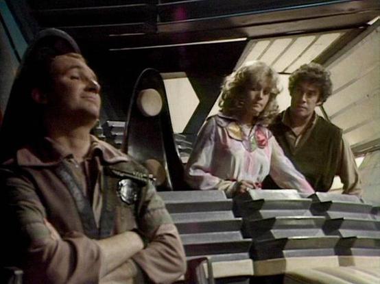Claude 3.7 describes the image as: "The image shows a scene from a science fiction television series, appearing to be from the late 1970s or early 1980s based on the production style and aesthetic. The setting appears to be the interior of a spacecraft or control room with distinctive angular panels and technical elements visible.

Three people are shown in the frame - one person in a brown jacket positioned to the left, looking slightly upward, while two others stand at what appears to be a co…