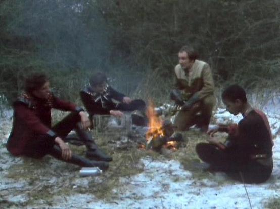 Claude Sonnet 4.0 describes the image as: "This scene shows four characters gathered around a small campfire in what appears to be a cave or rocky shelter. The setting suggests they're in hiding or taking refuge, with patches of snow or frost visible on the ground, indicating cold conditions. The characters are dressed in their typical practical clothing - leather jackets and sturdy garments suitable for their circumstances as rebels on the run. The intimate gathering around the fire creates a …