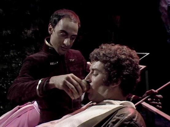 Claude 3.7 describes the image as: "The image shows a scene from a science fiction television series with a dark, moody atmosphere. Two people appear in what seems to be a futuristic setting. One person wearing a burgundy uniform is attending to another who has curly hair and appears to be lying down or injured. The standing figure is offering something to the person who is reclining.

The scene has dramatic lighting with dark shadows, typical of classic science fiction television production fr…