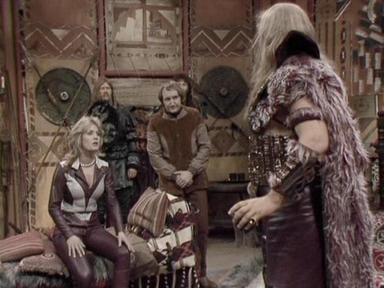 Claude 3.7 describes the image as: "This image shows a scene set in what appears to be a tribal or primitive interior space with decorative wall hangings and circular shields. Several people are visible in what looks like a costume drama from the late 1970s or early 1980s, based on the production style and costume design.

In the foreground, a person in leather-like attire sits on a cushioned seat, while others stand in the background. One figure wears a furry costume with an ornate vest, sugge…
