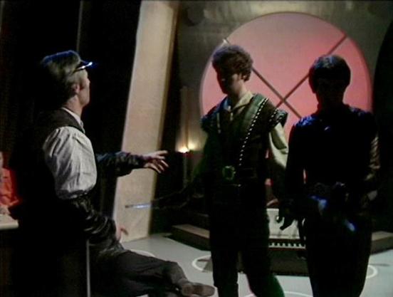Claude 3.7 describes the image as: "This image appears to be from a science fiction television production, showing three figures in a futuristic setting. The scene takes place in what looks like a spacecraft interior with distinctive lighting, including a circular red window or portal with an X pattern in the background.

The person on the left is seated and wearing period-style clothing with a white shirt, while gesturing toward the others. In the center stands someone in a green outfit with d…