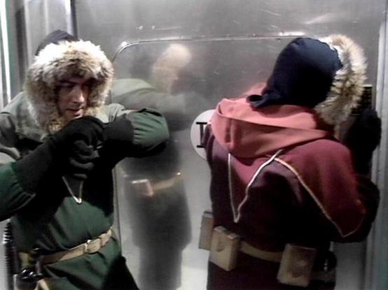 Claude 3.7 describes the image as: "The image shows two people in a scene that appears to be set in some kind of small enclosed space with metallic walls, possibly an elevator or small compartment. Both individuals are wearing winter clothing with fur-trimmed hoods. The person on the left wears a green jacket with gloves, while the person on the right is in a burgundy/red jacket with what looks like a utility belt. There's a reflection visible in what seems to be a glass or metal surface betwee…