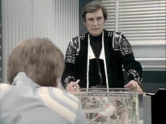Claude 3.7 describes the image as: "This image appears to be from a science fiction television series from the late 1970s or early 1980s. The scene shows two people in what looks like a futuristic setting with white walls or panels in the background.

In the foreground is a clear box or container that appears to contain some kind of mechanism or small object. The person standing is wearing a distinctive black costume with metallic shoulder pieces and embellishments - typical of the costume desi…