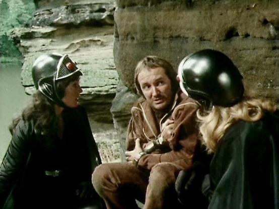 Claude 3.7 describes the image as: "This appears to be a scene from a classic science fiction television series from the late 1970s or early 1980s. The image shows three individuals in what looks like a rocky or cave-like setting. Two people are wearing distinctive black helmets with metallic finishes - one with a red sensor or light visible. The central figure is wearing a brown outfit and appears to be in conversation with or perhaps being interrogated by the helmet-wearing figures.

The prod…