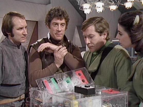 Claude 3.7 describes the image as: "This image appears to be from a science fiction television series from the late 1970s or early 1980s. The scene shows four people gathered around what looks like a clear control panel or device with colored buttons and components. They're in what appears to be a spacecraft or futuristic setting, indicated by the technical equipment and the overhead lighting fixtures.

The individuals are dressed in typical science fiction attire of that era - practical jumpsu…