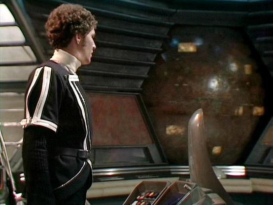 Claude Sonnet 4.0 describes the image as: "This image shows a scene set aboard a spaceship's bridge or control room. The setting features a futuristic interior with metallic walls, control panels, and what appears to be a large viewscreen or window showing space in the background. A person wearing a distinctive black outfit with white trim or piping is shown in profile, standing at what appears to be a control station or console. The costume design and set aesthetics are characteristic of late …