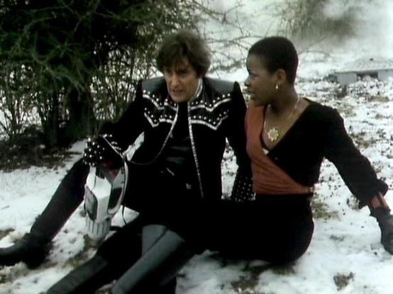 Claude Sonnet 4.0 describes the image as: "I can see two people sitting together in snow in what appears to be a winter outdoor setting. One person is wearing a distinctive black outfit with ornate white decorative trim and studs, while the other is dressed in dark clothing. They appear to be sitting close together on snowy ground with trees or foliage visible in the background. The scene has a dramatic, possibly romantic quality to it, and the winter setting creates an atmospheric backdrop for…