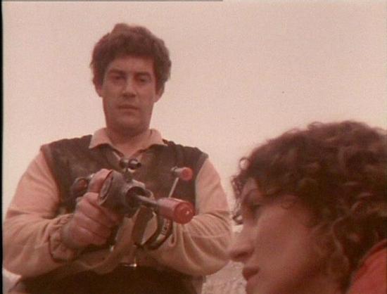 Claude 3.7 describes the image as: "This image appears to be from a vintage science fiction television series from the late 1970s or early 1980s. The still shows two people in what seems to be an outdoor setting with a light background. The person on the left is wearing a light-colored shirt with what looks like a leather vest or harness, and is holding what appears to be a prop weapon or futuristic gun with distinctive red elements. The production has that characteristic film quality of Britis…
