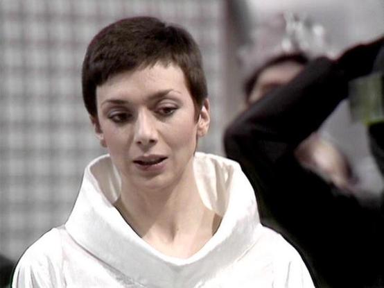 Claude 3.7 describes the image as: "The image shows a person with very short dark hair wearing a white robe or garment with a wide neckline. They have a focused expression and appear to be speaking or reacting to something. The background is blurry but suggests an indoor setting with some structural elements visible.

This appears to be from a science fiction production, with the minimalist costume and setting typical of the genre from that era. The stark white costume against the industrial-lo…
