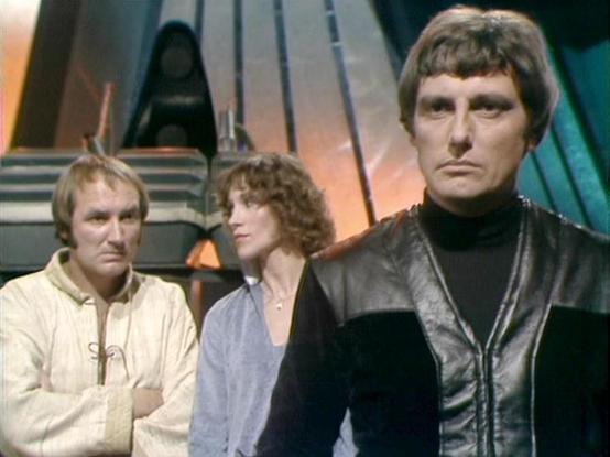 Claude Sonnet 4.0 describes the image as: "This appears to be a scene from a science fiction television series, showing three characters in what looks like a spaceship interior or futuristic setting. The scene has the distinctive visual style of late 1970s/early 1980s British sci-fi television, with metallic set pieces and atmospheric lighting in the background.
The three individuals are dressed in period-appropriate futuristic costumes - one in a light-colored tunic-style garment, another in …