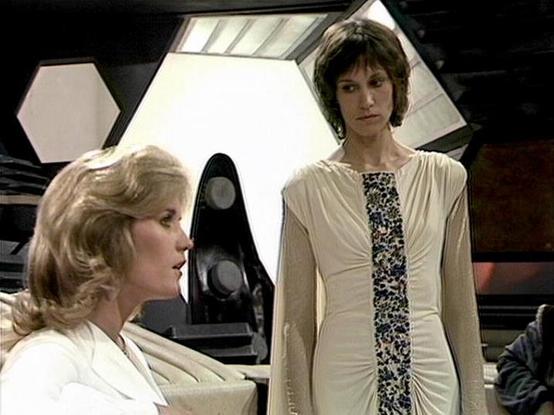 Claude 3.7 describes the image as: "This image appears to be from a science fiction television series, showing a scene set in what looks like a futuristic spacecraft interior. The setting features hexagonal wall panels and technical equipment in the background, creating a distinctive retro sci-fi aesthetic typical of productions from the late 1970s or early 1980s.

Two people are shown in conversation - one seated on the left wearing a white outfit with blonde hair, and another standing on the …