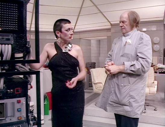 Claude 3.7 describes the image as: "This image appears to be from a science fiction television series, showing a scene set in a futuristic, sterile-looking interior space with white walls and minimalist design elements. 

The scene features two individuals in conversation. On the left is a person with very short dark hair wearing an elegant black one-shoulder dress and statement jewelry with a distinctive necklace. On the right is an older individual in a light gray or beige coat or lab coat.

…