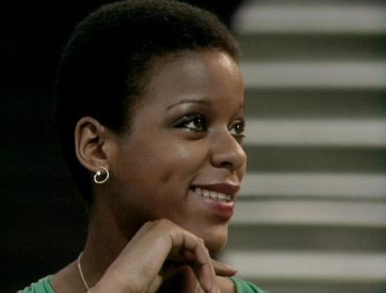 Claude Sonnet 4.0 describes the image as: "I can see this is an image of a woman with short dark hair wearing small hoop earrings and what appears to be a green or teal colored garment. She has her hand positioned near her chin in a thoughtful pose and is smiling at the camera. The background appears to show some kind of metallic or technological setting with horizontal striped elements, which would be consistent with a science fiction television production. The lighting and image quality sugge…