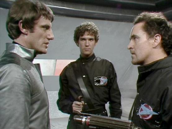 Claude Sonnet 4.0 describes the image as: "This scene appears to be set aboard a spacecraft, likely the Liberator, with its characteristic white and metallic interior design. Three crew members are engaged in what seems to be a tense discussion or confrontation in a corridor or command area of the ship. The characters are wearing the dark, fitted uniforms typical of the series, with visible insignia patches. The lighting and set design create the futuristic, somewhat sterile atmosphere characte…
