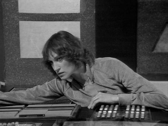 Claude Sonnet 4.0 describes the image as: "I can see this is a black and white image showing someone at what appears to be a control console or technical workstation. The person is leaning over the console, which has various buttons, switches, and control panels typical of science fiction television production design from the late 1970s or early 1980s. The setting appears to be some kind of spacecraft or high-tech facility interior, with geometric wall panels visible in the background. The ligh…