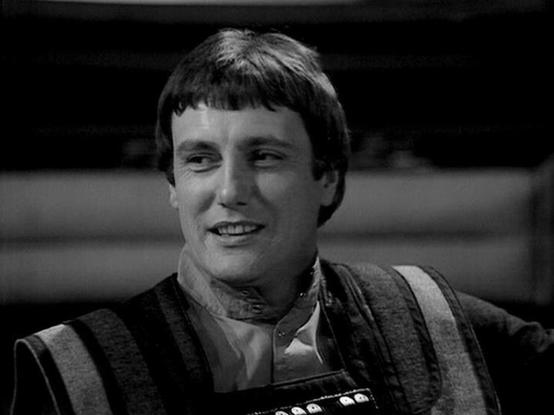 Claude 3.7 describes the image as: "This is a black and white image from a classic science fiction television series from the late 1970s/early 1980s. The image shows a person with short dark hair wearing what appears to be a distinctive costume with striped elements and a high collar, typical of the show's futuristic styling. 

The person has a slight smile and appears to be in what would be a spacecraft or base set from the series. The production values reflect the era of television in which i…