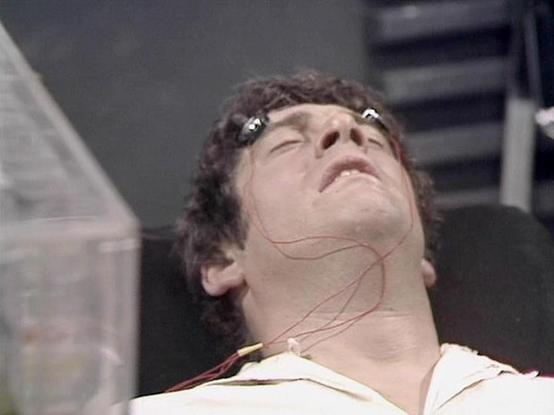 Claude 3.7 describes the image as: "The image shows a scene from a science fiction television production, appearing to be from the late 1970s or early 1980s based on the visual quality and style. In the frame, a person with dark curly hair is lying back with their eyes closed, appearing to be undergoing some kind of medical procedure or interrogation. There are thin red wires or electrodes attached to their face, suggesting some form of monitoring or mental probing device. They're wearing what …