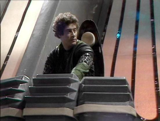 Claude 3.7 describes the image as: "This image appears to be from a science fiction television series from the late 1970s or early 1980s, based on the production style and set design. 

The scene shows a person with curly hair wearing a dark outfit with what looks like some kind of decorative or functional arm detail. They appear to be in a futuristic spacecraft or space station interior with distinctive angular design elements. In the foreground, there are what look like control panels or equi…