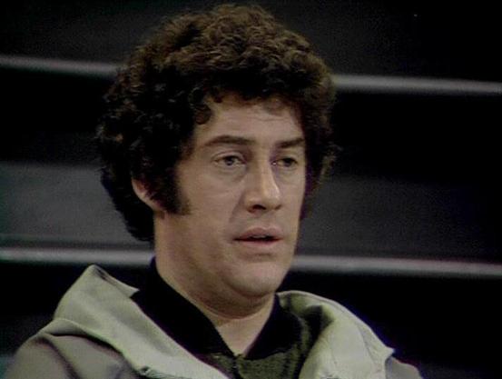 Claude 3.7 describes the image as: "The image appears to be from a classic science fiction television series from the late 1970s/early 1980s era, showing a person with curly dark hair wearing what looks like a light-colored vest or jacket over a darker garment. The background is dark, suggesting a spacecraft or futuristic setting typical of space-based sci-fi productions from that time period.

The styling, costume design, and filming quality are characteristic of British television productions…