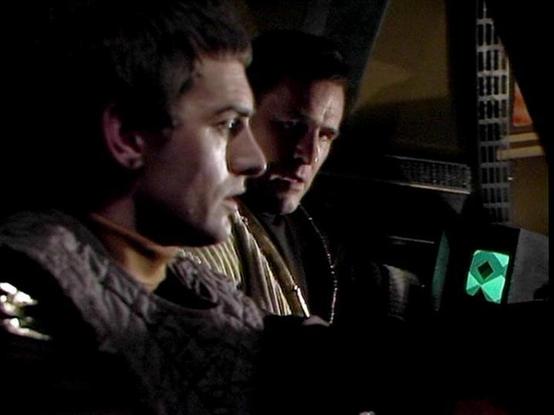 Claude Sonnet 4.0 describes the image as: "I can see two men in what appears to be the interior of a spacecraft or control room setting. They're positioned near what looks like control panels or technical equipment, with some kind of display screen visible showing a green geometric shape. The lighting is dramatic and moody, typical of science fiction television production from this era. The men are wearing what appears to be futuristic or military-style clothing. The setting suggests they're li…