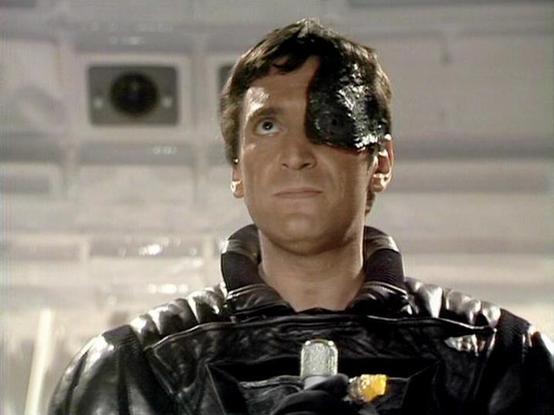 Claude 3.7 describes the image as: "The image shows a character from a science fiction television series wearing a distinctive black leather uniform with a silver badge or emblem on the front. The character has a notable eye patch or covering over one eye, which is a defining feature of their appearance. This appears to be from a scene set on a spaceship or futuristic facility, as indicated by the sterile, technological-looking background.

The costume design is characteristic of dystopian sci-…