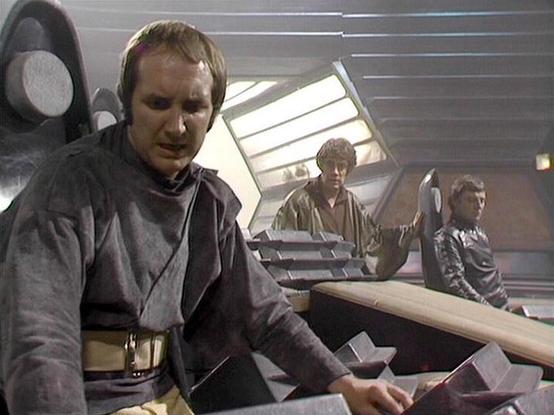 Claude 3.7 describes the image as: "This image shows a scene from what appears to be a science fiction television production from the late 1970s or early 1980s, based on the visual style and production quality. The setting appears to be the bridge or control room of a spacecraft, with futuristic control panels and a distinctive angular design aesthetic typical of sci-fi productions from that era.

Several people are visible in the control room, wearing costume garments that suggest a unified st…