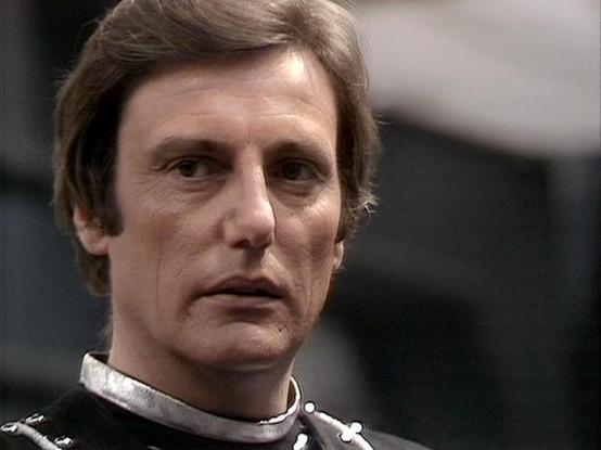 Claude Sonnet 4.0 describes the image as: "This image shows a character in what appears to be a futuristic military or space-faring uniform, featuring a high collar with metallic detailing. The costume design reflects the science fiction aesthetic typical of late 1970s British television production. The setting appears to be an interior spaceship or facility environment with blurred background elements. The uniform's sleek, authoritarian styling with its reflective collar elements suggests this…