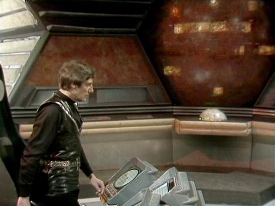 Claude 3.7 describes the image as: "This image shows a scene from a science fiction television series, set in what appears to be a spaceship or control room. The setting has distinctive angular walls with reddish-brown paneling and hexagonal design elements typical of 1970s/80s sci-fi productions.

In the scene, a person wearing a dark leather outfit stands at what looks like a control console with various technical equipment. The futuristic control panel contains box-like devices with displays…