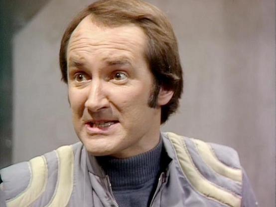 Claude 3.7 describes the image as: "The image shows a person with brown hair wearing a gray and cream-colored vest over a dark turtleneck. They appear to be in a scene from a science fiction television production from the late 1970s or early 1980s, as suggested by the distinctive costume styling and visual quality of the footage.

The individual has an expressive facial expression showing concern or mild distress. The background appears minimal and somewhat sterile, consistent with spacecraft o…