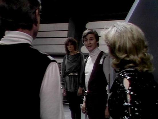 Claude 3.7 describes the image as: "The image shows a scene from a science fiction television series from the late 1970s/early 1980s. It appears to be set in a futuristic corridor or spacecraft interior with clean, minimalist design elements typical of that era's sci-fi aesthetic.

The scene captures several people in conversation, wearing distinctive costumes that reflect the show's styling. One person is dressed in a gray draped outfit, while another wears a white turtleneck with a dark vest.…