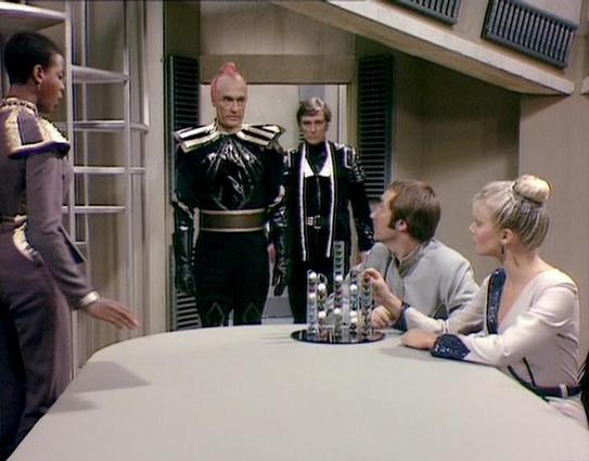 Claude 3.7 describes the image as: "This image shows a scene set in what appears to be a futuristic space station or spacecraft interior with a minimalist design aesthetic typical of science fiction from the late 1970s/early 1980s. 

The scene features several people in distinctive costumes around a curved white table that contains what looks like a small display or game board with colored pieces. Two figures stand wearing dramatic black uniforms with striking shoulder details, conveying author…