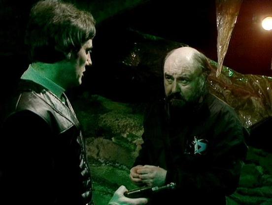 Claude 3.7 describes the image as: "This image shows a scene set in what appears to be a dark, cavernous environment with a greenish tint to the lighting. Two people are engaged in conversation - one younger figure on the left wearing a leather-type jacket and another older figure with a beard on the right in dark clothing with what looks like an emblem or badge. The setting suggests an underground or secluded location, typical of the science fiction aesthetic from television productions of the…