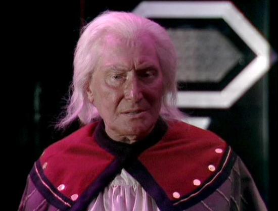 Claude 3.7 describes the image as: "This image appears to be from a science fiction television production, featuring a person with distinctive white/pink hair wearing an elaborate costume. They're dressed in a red and purple outfit with decorative elements including what looks like buttons or studs along the collar. 

The setting has a futuristic feel with what appears to be geometric shapes or control panels visible in the background, typical of science fiction set design from the late 1970s o…