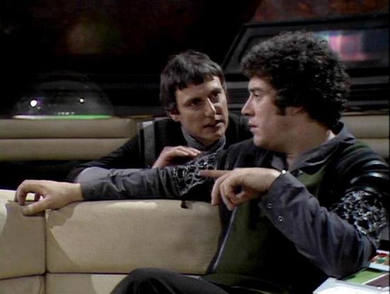 Claude 3.7 describes the image as: "The image shows two people in what appears to be a spacecraft interior, sitting on cream-colored seating. They're engaged in what seems to be a serious conversation. Both are wearing dark clothing with some decorative elements - one has curly hair while the other has straight darker hair. The futuristic setting is suggested by the control panel visible in the background with colored lights. The aesthetic has that distinctive late 70s/early 80s sci-fi televisi…
