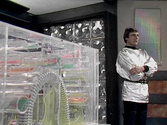 Claude 3.7 describes the image as: "This image shows a scene from a science fiction television series set in what appears to be a futuristic control room or laboratory. In the foreground is a transparent or clear equipment panel with various colorful components visible inside. The background features metallic wall panels with a distinctive textured design.

A person in a white tunic-style uniform with crossed arms is standing to the right side of the image, positioned near what looks like a doo…