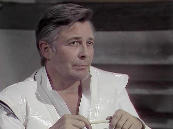 Claude Sonnet 4.0 describes the image as: "I can see this appears to be from a science fiction television production, likely from the late 1970s or early 1980s based on the visual style and production values. The scene shows someone in what appears to be a futuristic white outfit or uniform, sitting in what looks like a spaceship or futuristic setting. The lighting and set design have that distinctive look characteristic of British sci-fi productions from that era, with clean, minimalist aesthe…