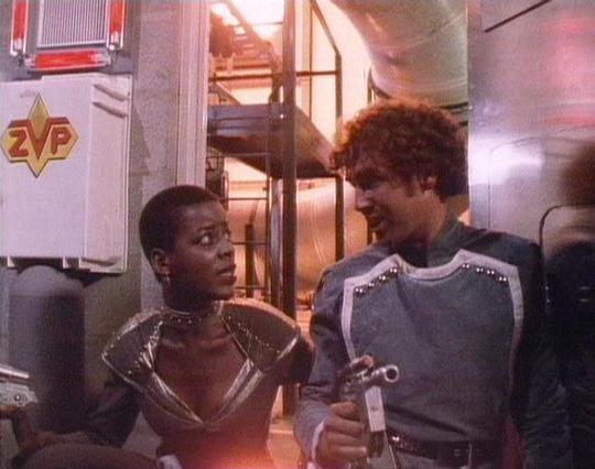 Claude Sonnet 4.0 describes the image as: "I can see this appears to be from a science fiction television series, showing two people in what looks like a spacecraft or futuristic setting. The scene takes place in what appears to be a corridor or room with metallic walls and technological equipment visible in the background. One person is wearing what appears to be a form-fitting outfit with metallic or reflective elements, while the other is dressed in what looks like a light-colored uniform or…