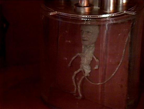 Claude Sonnet 4.0 describes the image as: "I can see this appears to be a science fiction scene featuring what looks like a transparent cylindrical chamber or containment unit with a reddish tint. There appears to be a figure visible within the chamber, though the image quality and lighting make details difficult to discern clearly. The setting has a futuristic, clinical appearance typical of sci-fi television production design from this era, with metallic elements visible at the top of the cha…