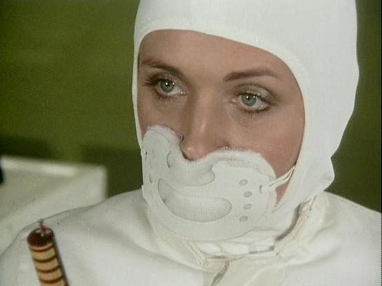 Claude Sonnet 4.0 describes the image as: "I can see this image shows someone wearing a white protective suit or uniform with a distinctive helmet or head covering that has breathing apparatus around the mouth area. The costume appears to be from a science fiction production, with a clean, clinical aesthetic typical of futuristic medical or hazmat gear. The setting appears to have a greenish background, suggesting it might be from a medical facility or laboratory scene. The protective gear cove…