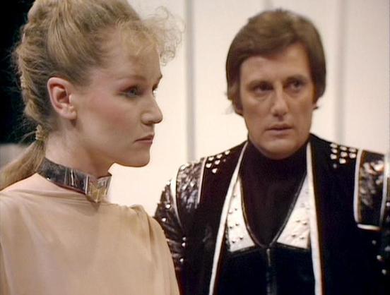 Claude Sonnet 4.0 describes the image as: "I can see two people in what appears to be a futuristic setting with white/light colored walls or corridors typical of science fiction productions from this era. One person is wearing an ornate costume with metallic studs and decorative elements in black and white, suggesting a military or authoritative role. The other person is wearing a simpler light-colored garment with what appears to be a collar or neck piece. The lighting and set design have the …