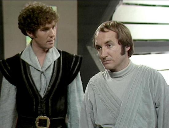 Claude 3.7 describes the image as: "The image shows a scene from a science fiction television production with a distinctive 1970s-80s aesthetic. Two performers are pictured in costume - one wearing a black and white medieval-inspired outfit with a belt, while the other is dressed in a simple light-colored tunic or jumpsuit with textured fabric.

The setting appears to be inside a futuristic spacecraft or base with louvered panels visible in the background. The lighting and film quality have the…