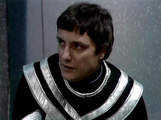 Claude Sonnet 4.0 describes the image as: "This image shows a scene from what appears to be a science fiction television production, featuring a character in distinctive futuristic costume. The person is wearing a striking black outfit with metallic silver trim arranged in V-shaped patterns across the chest and shoulders. The costume has a high collar and appears to be made of leather or similar material, giving it a sleek, space-age appearance typical of 1970s-80s sci-fi productions.

The sett…