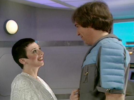 Claude 3.7 describes the image as: "The image shows two people in what appears to be a futuristic spaceship or station interior. One person with very short dark hair is wearing a light-colored patterned outfit and is looking up with a smile at the other person, who is wearing a distinctive blue padded vest or jacket with gray sections. 

The setting has a clean, minimalist sci-fi aesthetic typical of classic British television productions, with rounded wall fixtures and technical-looking panels…