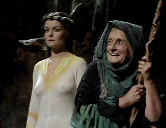 Claude Sonnet 4.0 describes the image as: "I can see two figures in what appears to be a dramatic scene. One person is wearing a light-colored, flowing garment and has an ornate headpiece or crown. The other figure is dressed in dark, hooded robes and is holding what looks like a wooden staff or walking stick. The lighting and staging suggest this is from a television production, with dramatic shadows and atmospheric lighting typical of science fiction or fantasy programming from that era. The …