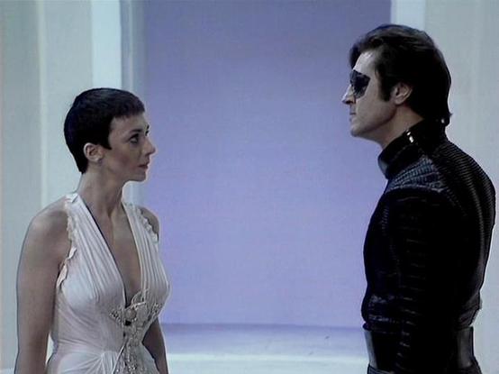 Claude Sonnet 4.0 describes the image as: "I can see two people in what appears to be a futuristic sci-fi setting with distinctive lighting and architectural elements typical of the series. One person is wearing an elegant white dress with decorative details, while the other is dressed in dark leather or similar material with sunglasses, creating a striking contrast between their costumes. The scene has a dramatic, tense quality to it, with the lighting creating an otherworldly atmosphere. The …